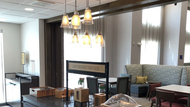 A modern dining area with a long table, hanging pendant lights, and a sign indicating breakfast service.