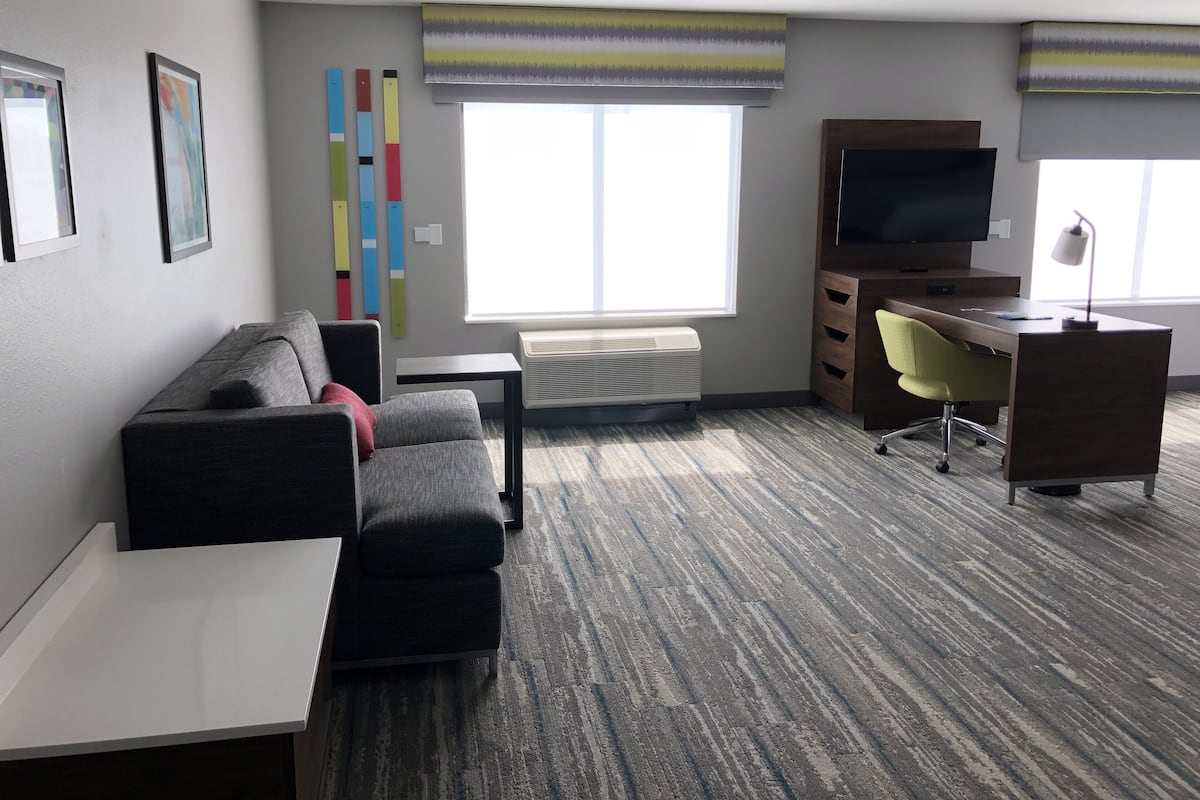 A modern hotel room with a gray sofa, a small table, a desk with a chair, a TV, and a window with a striped curtain.