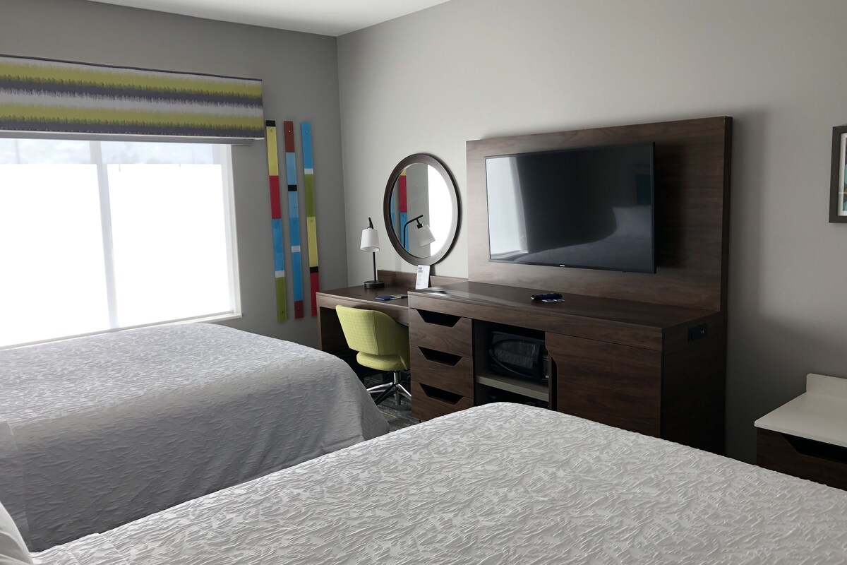 Une chambre d’hôtel avec deux lits, un bureau avec une télévision, un miroir et une fenêtre avec des stores.