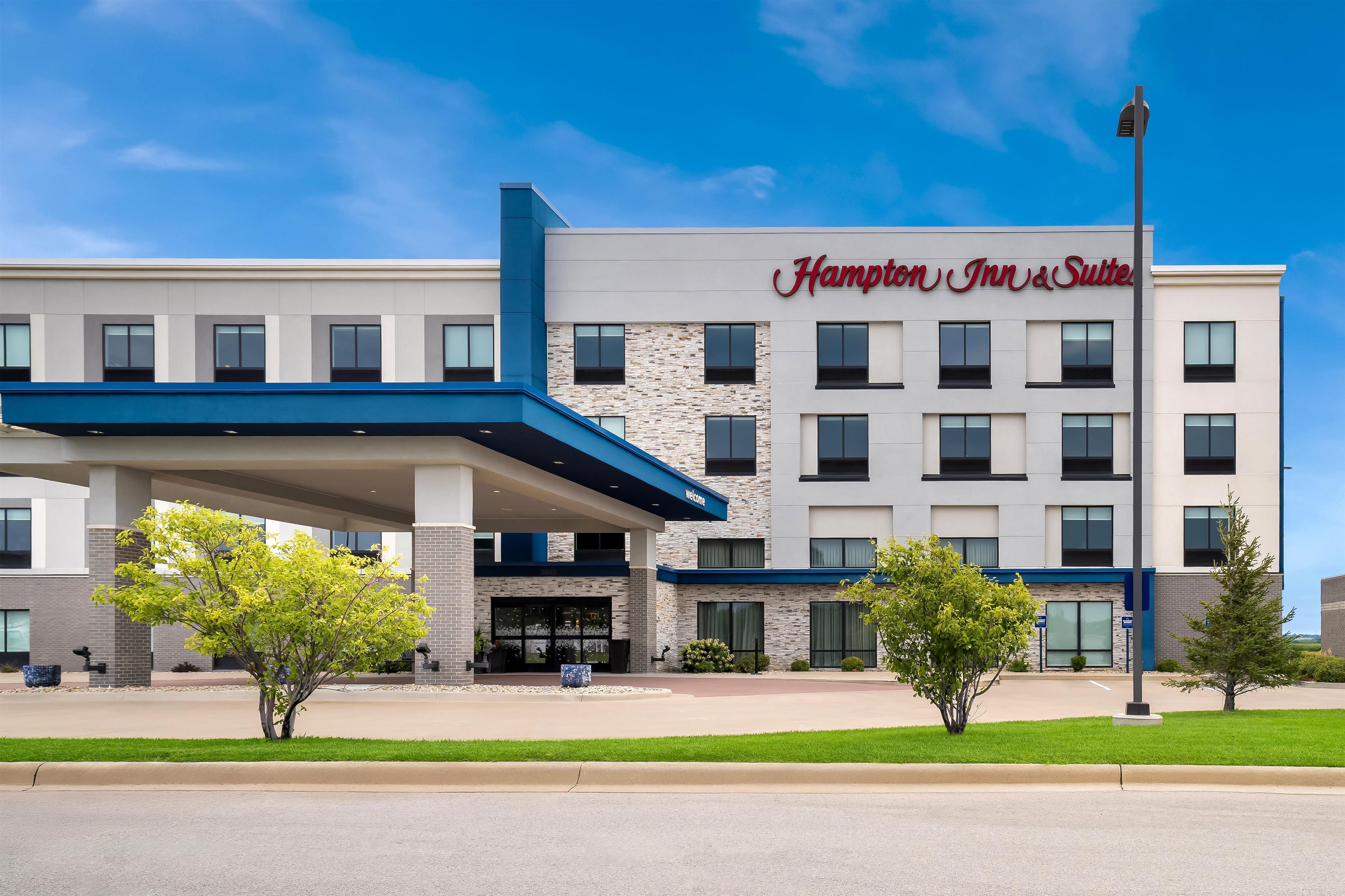Un hôtel Hampton Inn & Suites au design moderne, avec une entrée imposante et un ciel d’un bleu éclatant.