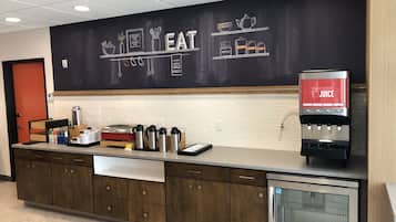Un área de cocina moderna con una pared de pizarra que muestra la palabra "EAT" y varios utensilios de cocina. Hay un dispensador de jugo y un refrigerador.