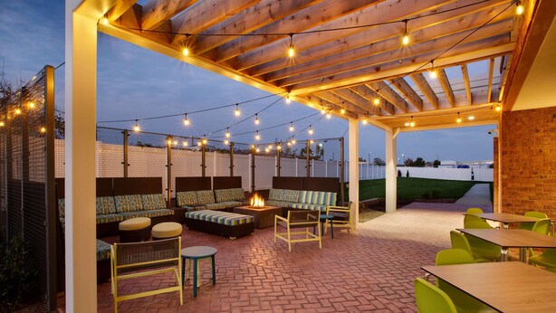 An outdoor patio area with a wooden pergola, string lights, a fire pit, seating, and tables. - Home2 Suites by Hilton Lewes Rehoboth Beach (Lewes)