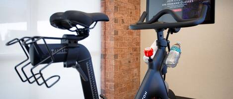 A Peloton stationary bike with a screen displaying a class advertisement, a water bottle holder, and a kickstand.