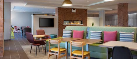 A modern restaurant with a brick wall, a flat-screen TV, and a fireplace. There are wooden chairs and tables, and a green sofa with colorful cushions.