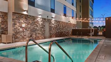A swimming pool with stone walls and metal railings, surrounded by a building with large windows and string lights.