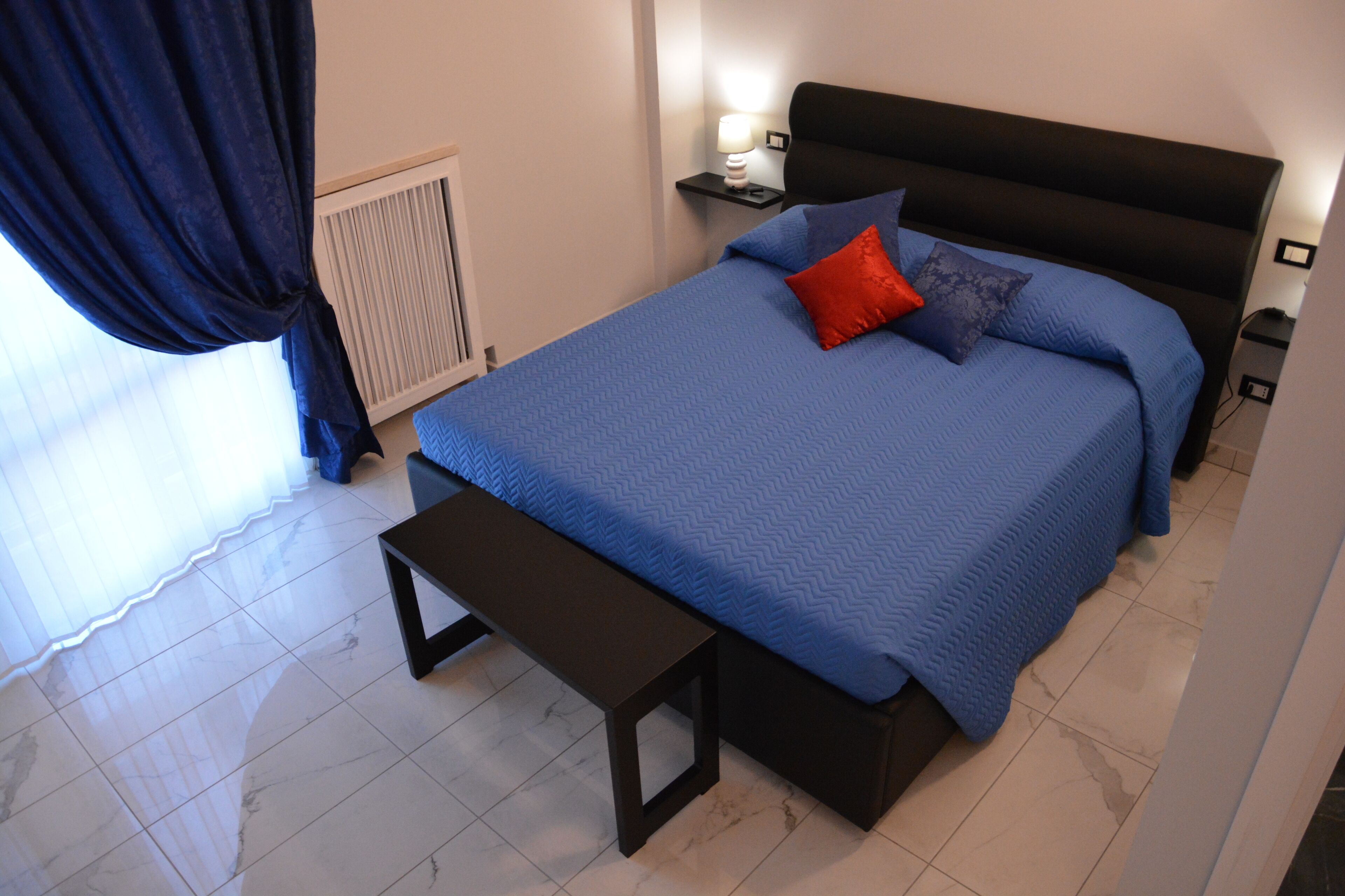 A bedroom with a bed, a bedside table, a nightstand, a lamp, and a window with curtains.