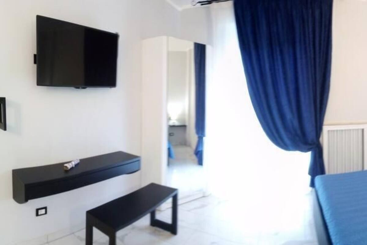 A bedroom with a bed, a bench, a TV mounted on the wall, and a window with curtains.