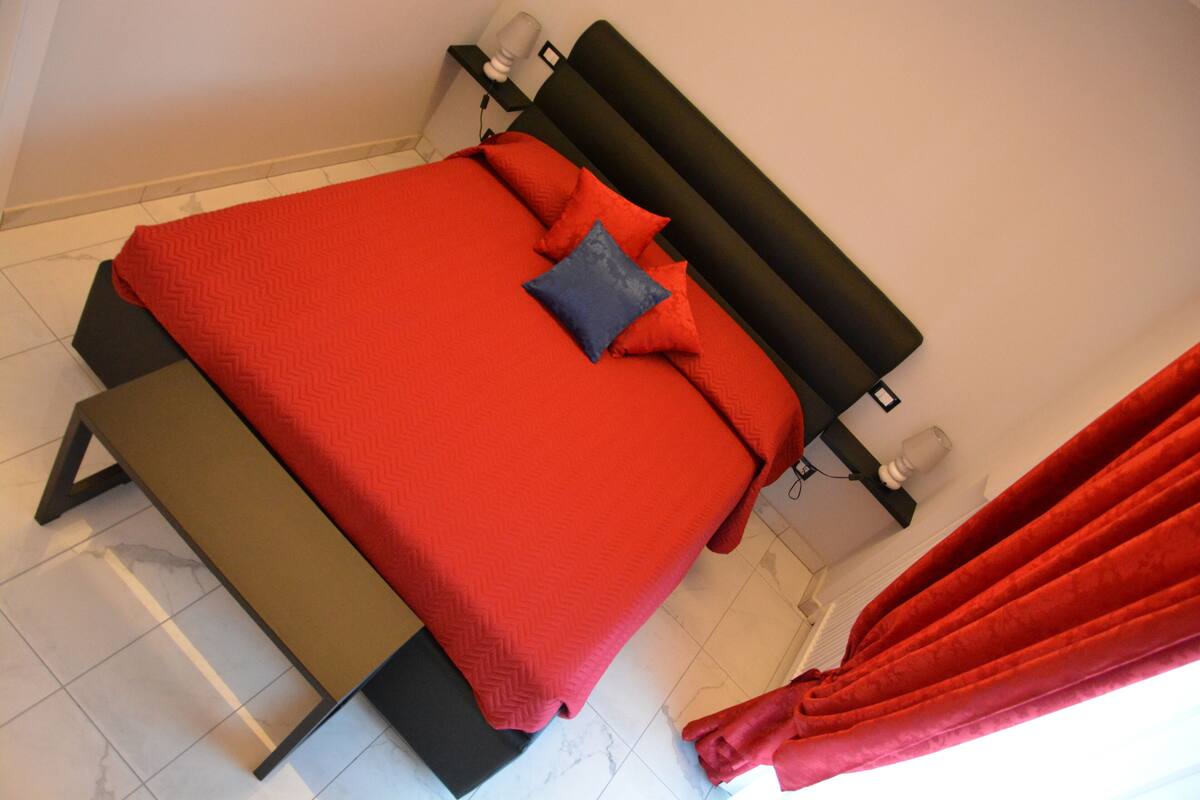 A bed with a red bedspread and pillows, a black headboard, and a red curtain.