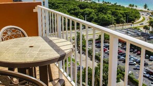 Un balcón con una mesa y sillas, con vista a un estacionamiento y una playa con palmeras y el océano.