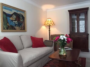 A living room with a beige sofa, red cushions, a table with a vase of flowers, a lamp, and a cabinet with a view of the water. - The Blue House (Westport)