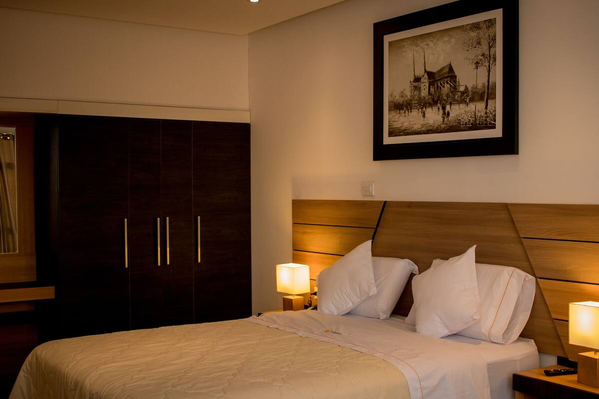 Ein Hotelzimmer mit einem großen Bett, einem Holz-Kopfteil, zwei Nachttischlampen, einem gerahmten Bild an der Wand und einem dunklen Holzschrank.