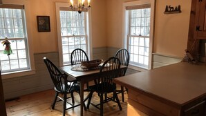 A cozy kitchen with wooden floors, a dining table with chairs, a chandelier, and large windows with a view of trees. - Cozy cottage tucked away in the woods (Townshend)