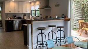 A modern kitchen with white cabinets, a central island with stools, and a dining area with a wooden table and blue chairs. - Pristine home w/heated saltwater pool, just minutes from beaches and bike trail. (Eastham)