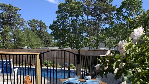 A patio with a pool, surrounded by a wooden fence, featuring a table and chairs, and blooming flowers. - Pristine home w/heated saltwater pool, just minutes from beaches and bike trail. (Eastham)