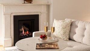 A cozy living room with a fireplace, a round coffee table with a book and two glasses of champagne, and a plush sofa with patterned cushions. - King with Kitchenette and Fireplace, Private Bath, Parking, Walk to all (Newport)