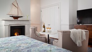 A bedroom with a fireplace, a sailboat decoration, a bed with a patterned bedspread, a small round table with glasses and a bottle, and a television on the wall. - King with Kitchenette and Fireplace, Private Bath, Parking, Walk to all (Newport)