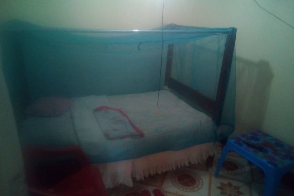 Una habitació petita i poc il·luminada amb un llit sol cobert per una mosquiterola. Es veu un petit tamboret i un gaster.