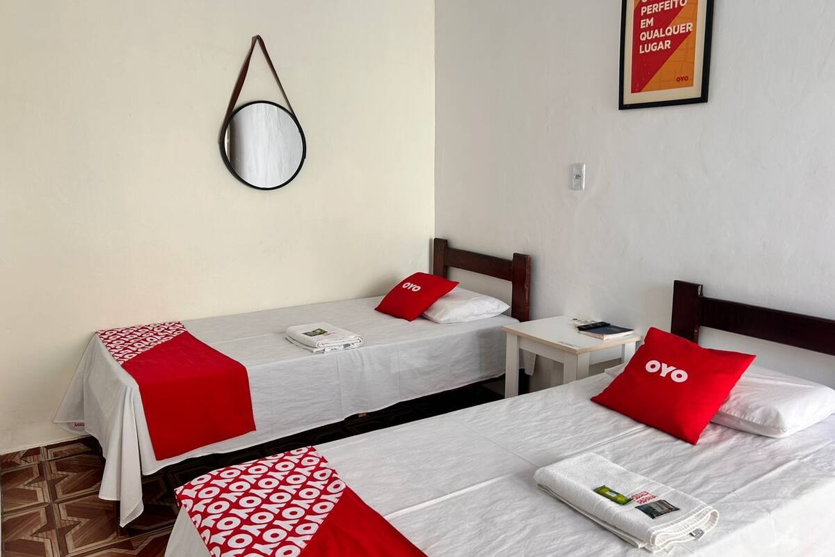 A hotel room with two beds, each with a red OYO branded pillow and a white bedspread. There is a small table between the beds, and a framed poster on the wall.