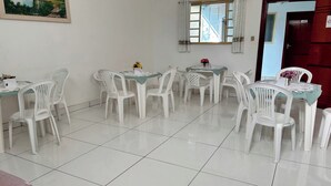 A dining area with white plastic chairs and tables, a painting on the wall, a window with curtains, and a door with a sign. - OYO Pousada Paraná, Cuiabá (Cuiabá)