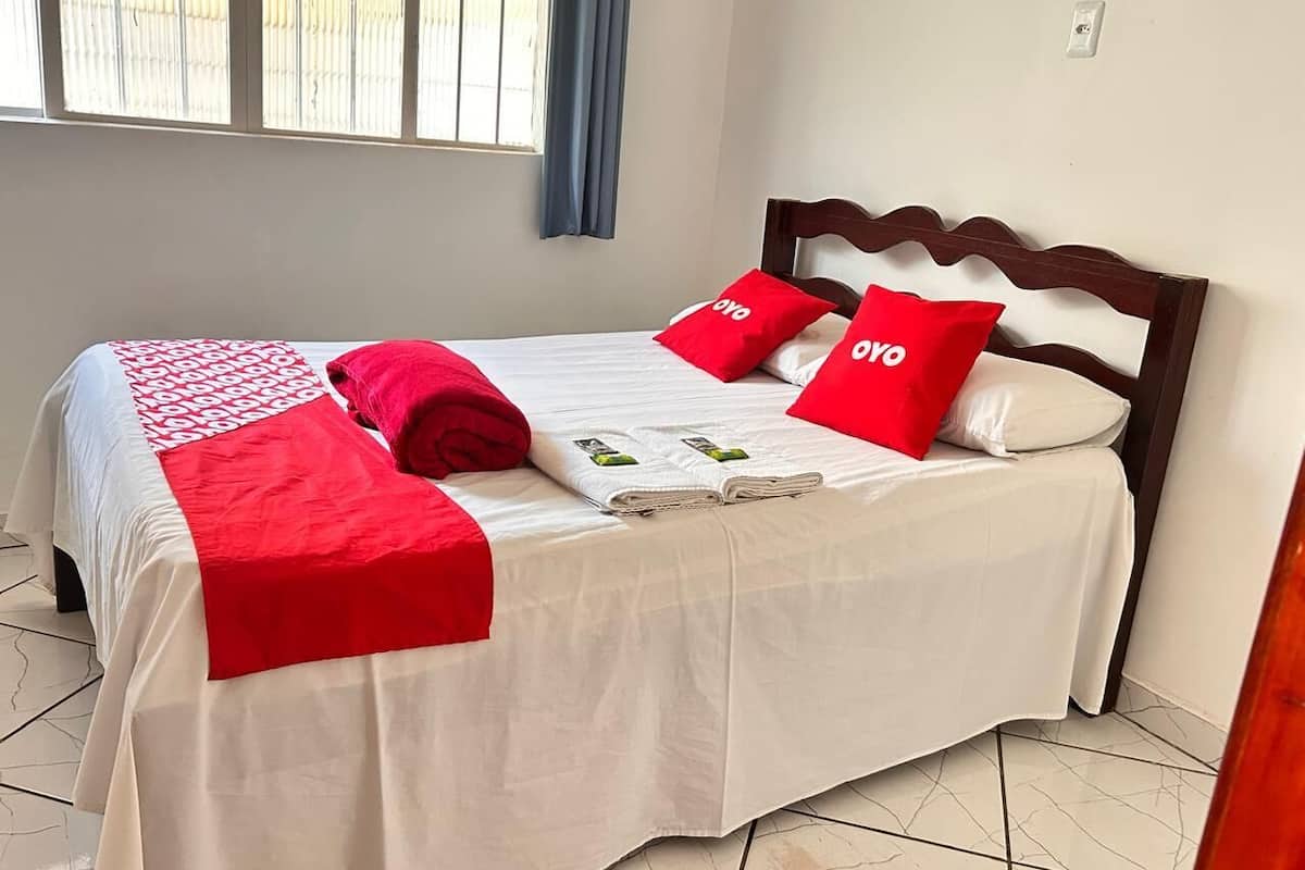 A neatly made bed with white and red bedding, a wooden headboard, and a window with blinds.