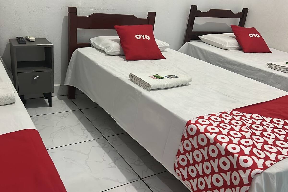 A hotel room with two single beds, each with a red pillow and a white bedspread. There is a small bedside table with a remote control and a book. The room has white tiled flooring.