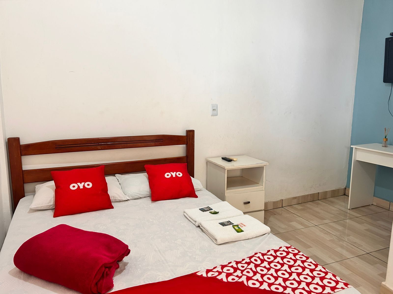 A neatly made bed with red and white bedding, a wooden headboard, a bedside table with a remote control, and a small white cabinet.