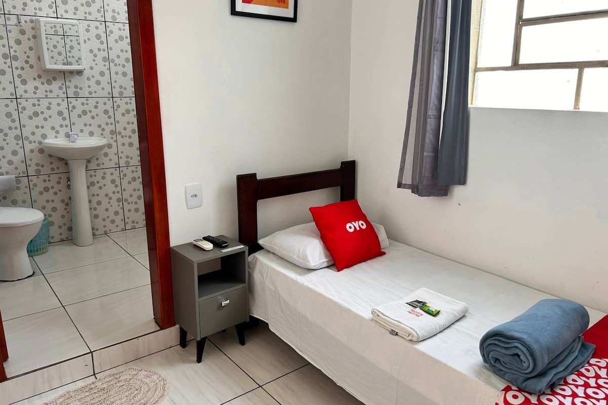 A small, neatly arranged bedroom with a single bed, a bedside table, a nightstand, a white sink, and a window with curtains.
