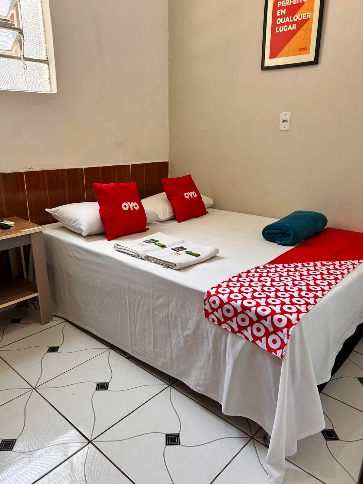 A hotel room with a bed, a nightstand, a red and white patterned blanket, and a poster on the wall.
