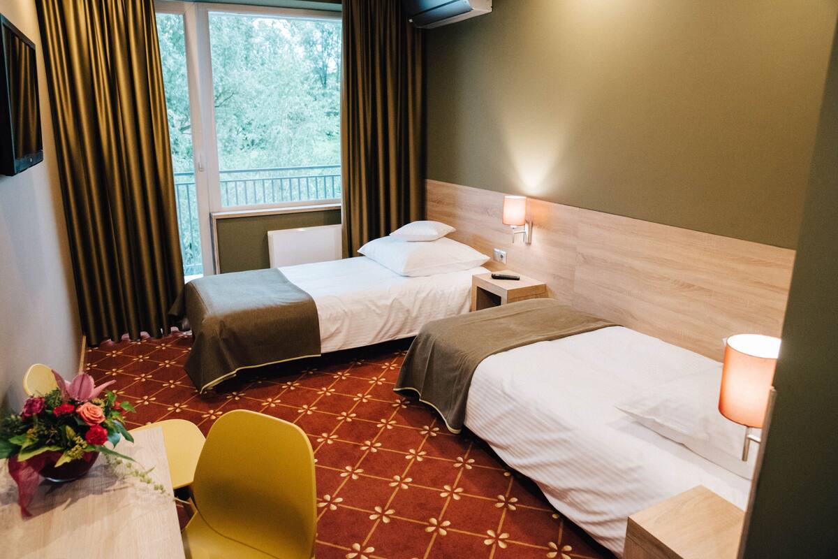 Habitación de hotel con dos camas, un escritorio, una silla, un florero con flores y una ventana con cortinas.