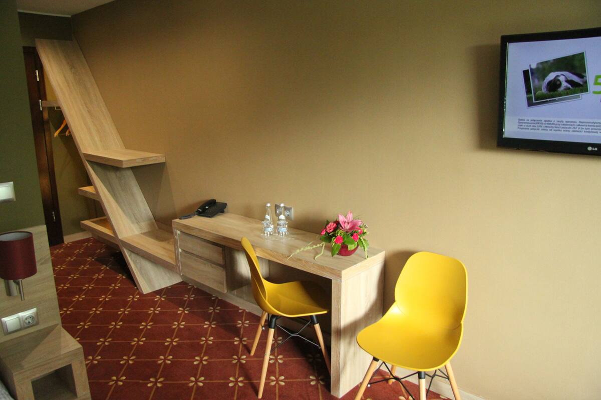 Una habitación con un escritorio, dos sillas amarillas, una mesita con un jarrón de flores, un teléfono y un televisor montado en la pared.