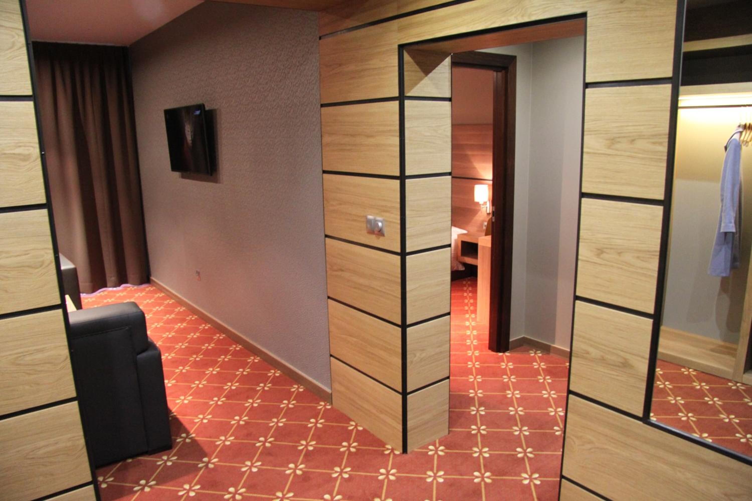 Een hotelkamer met een rood kamerbreed tapijt, houten paneelwanden, een zwarte zithoek en een spiegel die een deel van de kamer weerkaatst.