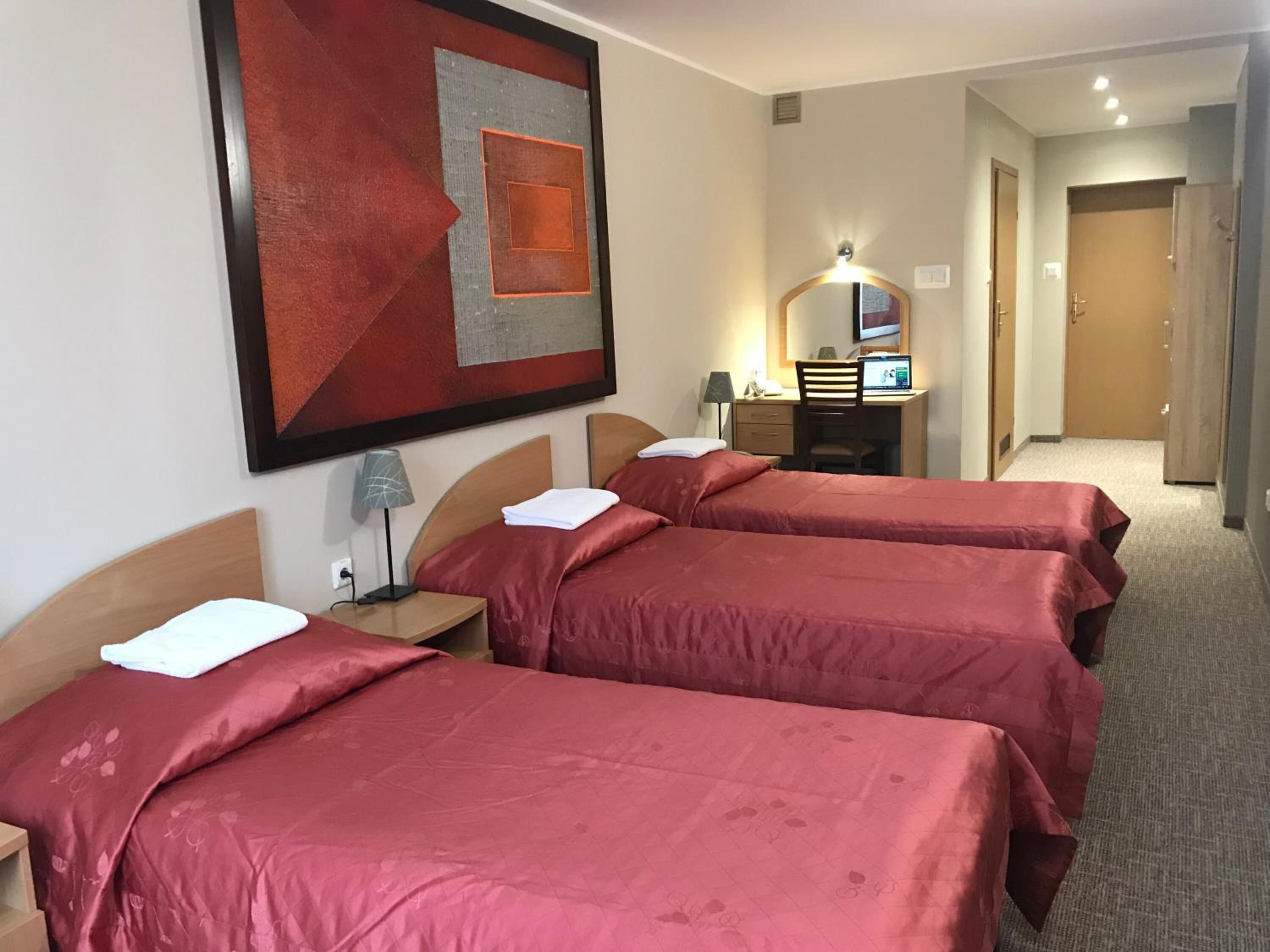 A hotel room with three beds, a desk with a computer, a mirror, and a large abstract painting on the wall.