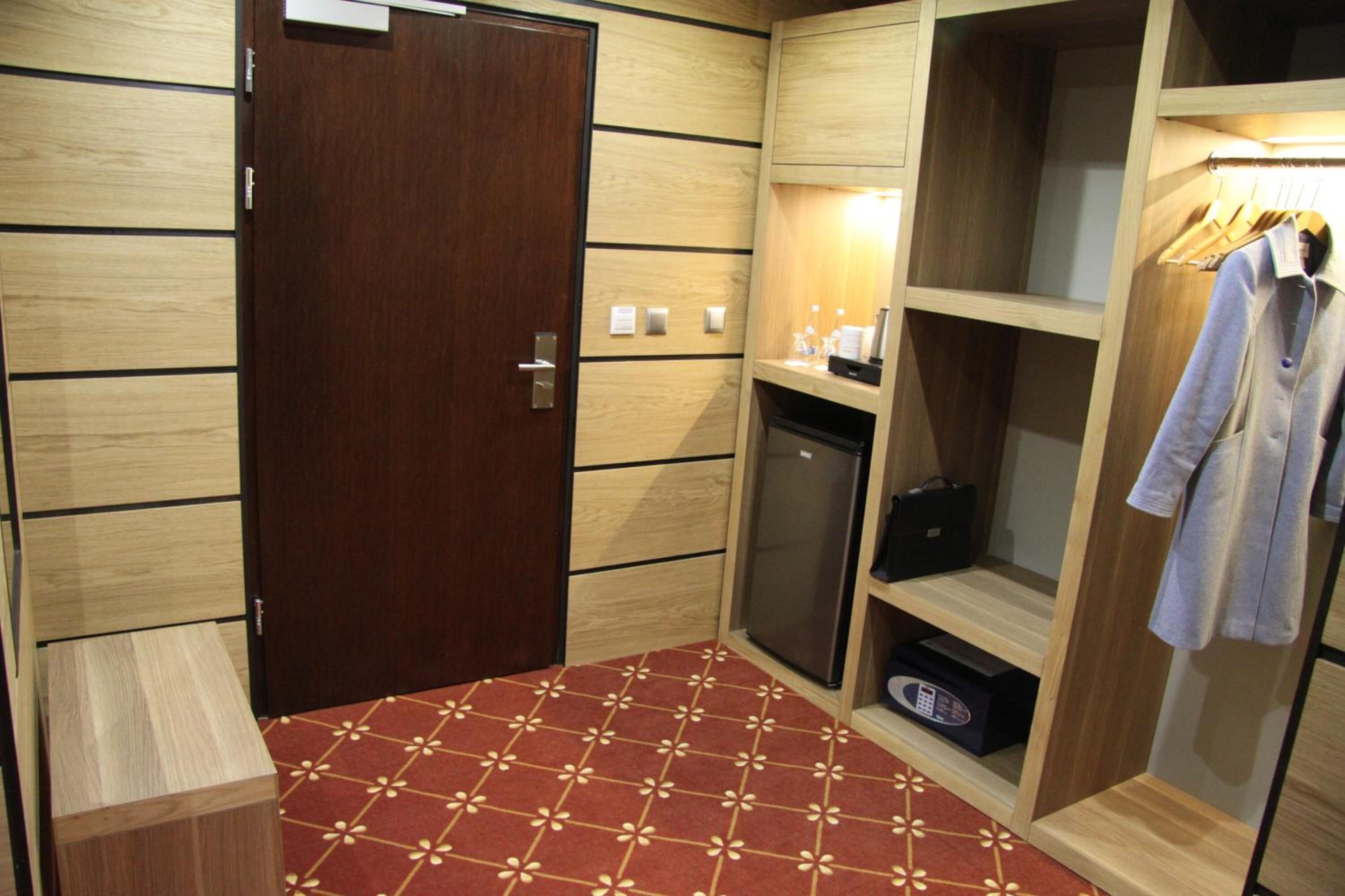 Ein Hotelzimmer mit Holzschrank, kleinem Kühlschrank, Spiegel und einem roten Teppich mit Muster.