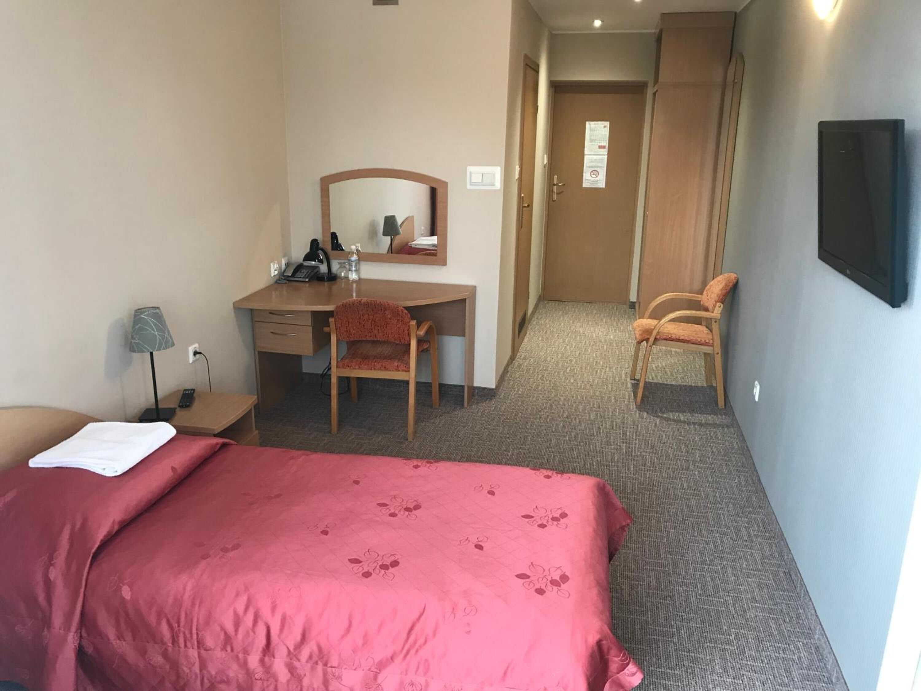 Habitación de hotel con cama, escritorio con silla, espejo, televisor montado en la pared y puerta que da a un pasillo.