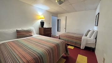A hotel room with two beds, a nightstand, a lamp, and a door leading to another room.