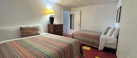 Habitación de hotel con dos camas, una mesita de noche, una lámpara y una puerta que da a otra habitación.