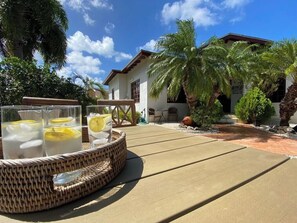 Un patio con una mesa de fibras naturales que sostiene vasos con una bebida y rodajas de limón, una casa con techo rojo y palmeras al fondo.