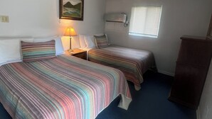 A room with two beds, a nightstand with a lamp, a window with blinds, a wall-mounted heater, and a painting.