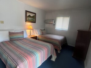 A room with two beds, a nightstand with a lamp, a window with blinds, a wall-mounted heater, and a painting. - Lakefront Suite - 14 (Kingsland)