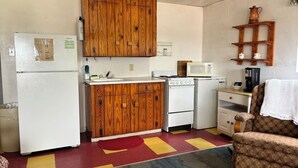 A compact kitchen with a refrigerator, stove, microwave, wooden cabinets, and a chair.