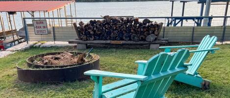 A lakeside outdoor seating area with a fire pit, a pile of firewood, and a sign indicating a burn ban.