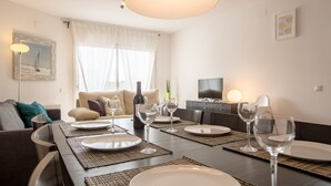 Ein Essbereich mit Tisch für vier Personen, komplett mit Weingläsern und Tellern, in einer modernen Wohnung, im Hintergrund ein Sofa und ein Fernseher.