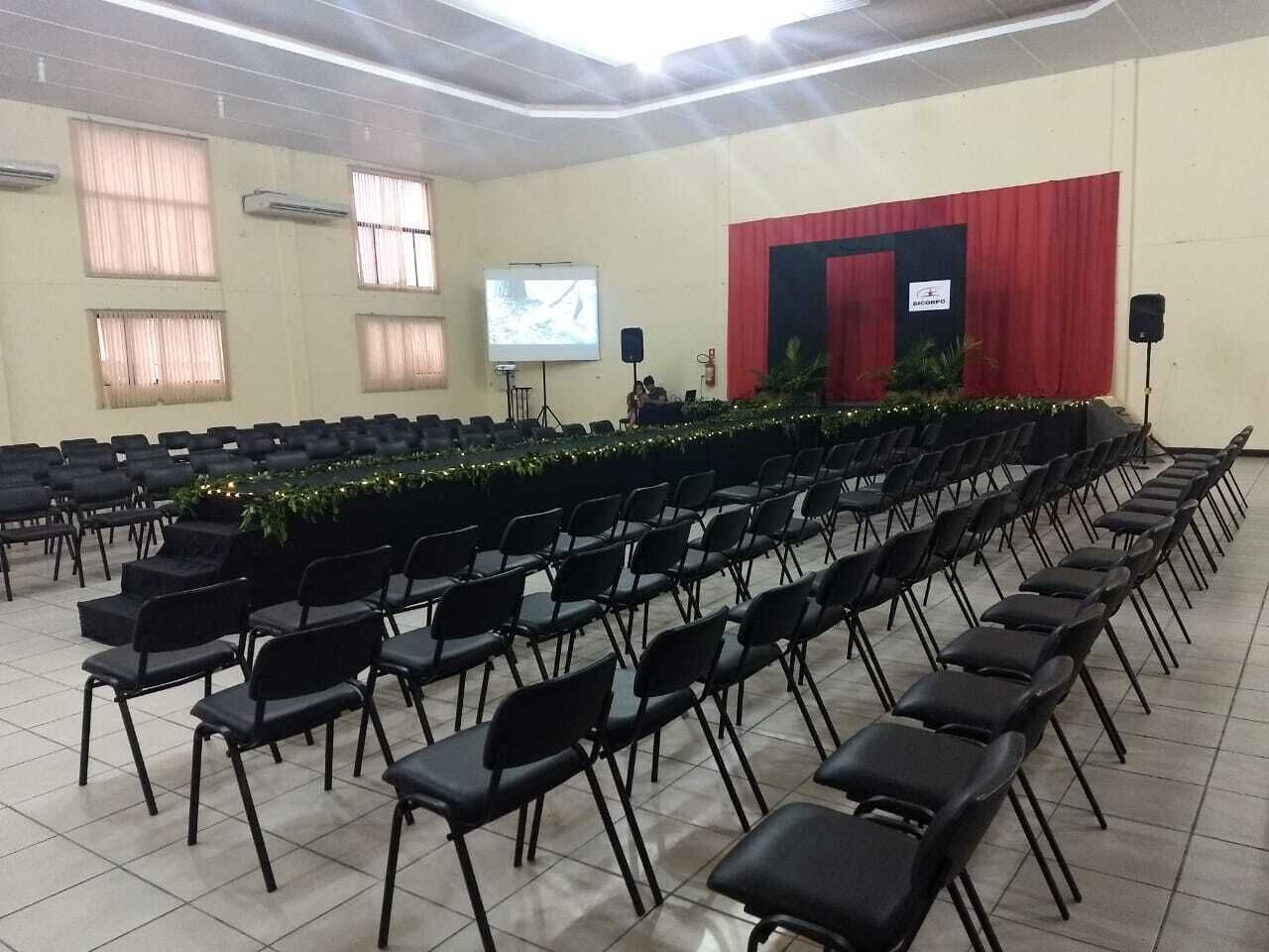 Un salón de conferencias con hileras de sillas negras, un escenario con cortina roja, una pantalla de proyector y un orador.