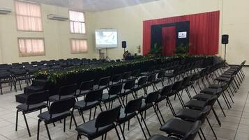Un salón de conferencias con hileras de sillas negras, un escenario con cortina roja, una pantalla de proyector y un orador.
