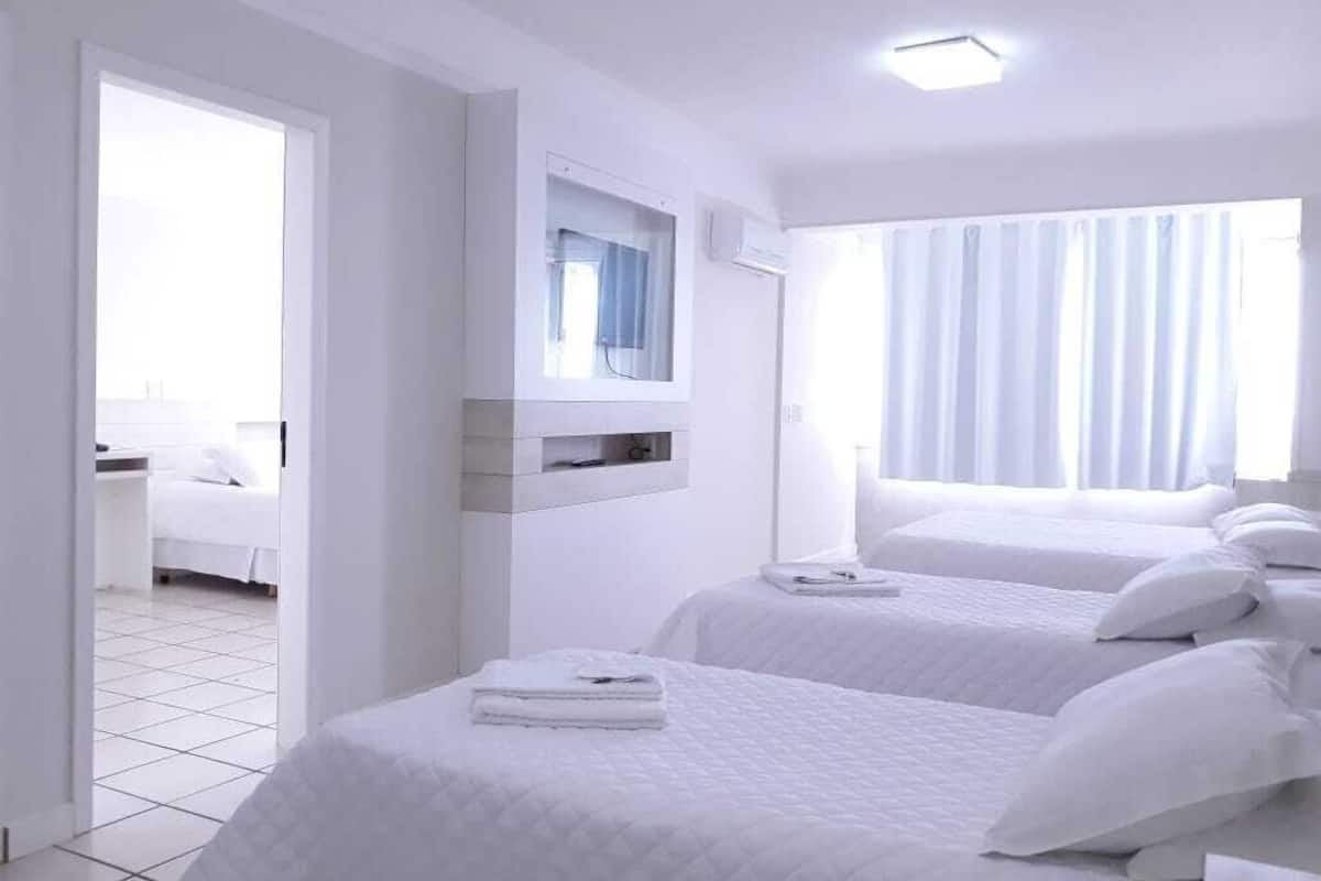 Ett hotellrum med två sängar, ett fönster med gardiner, en inbyggd hylla och en dörr som leder till ett annat rum.