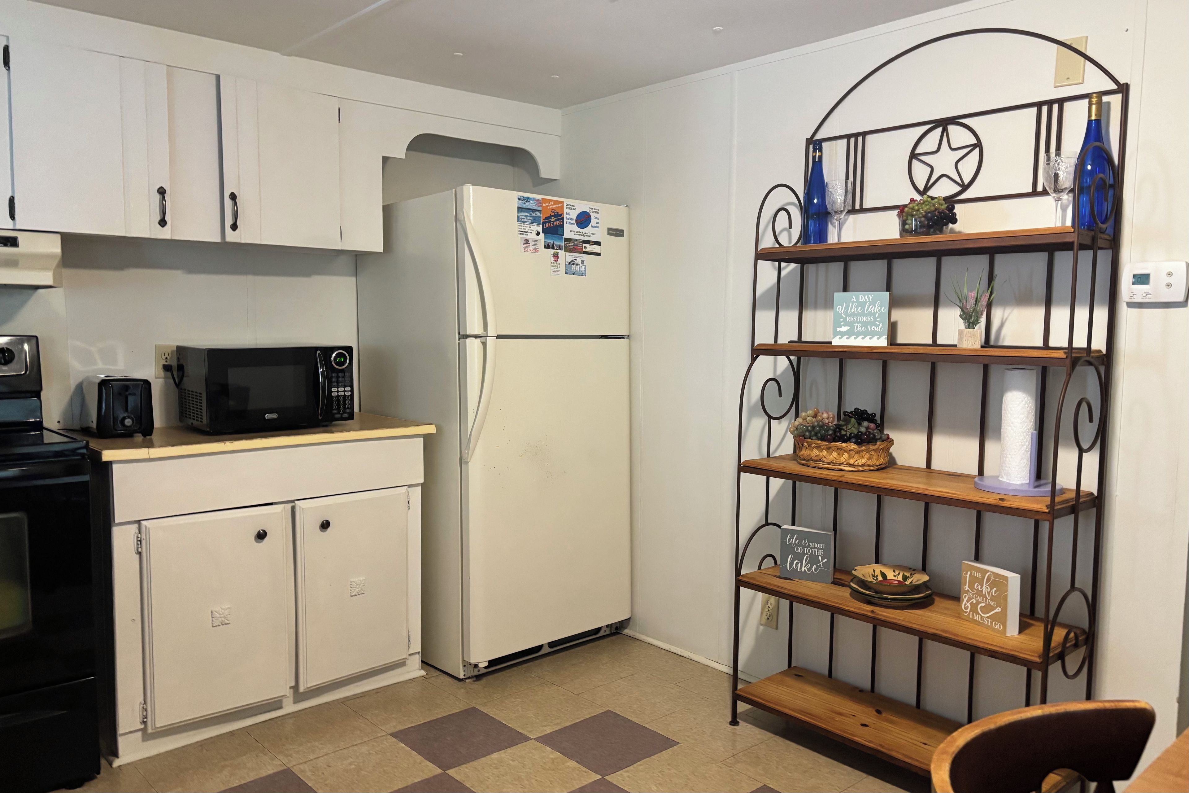 A compact kitchen with white cabinets, a white refrigerator, a microwave, a toaster, and a small stove. There is a metal shelf with decorative items and a sign that reads 'A day at the lake restores the soul.'