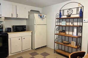 A compact kitchen with white cabinets, a white refrigerator, a microwave, a toaster, and a small stove. There is a metal shelf with decorative items and a sign that reads 'A day at the lake restores the soul.'