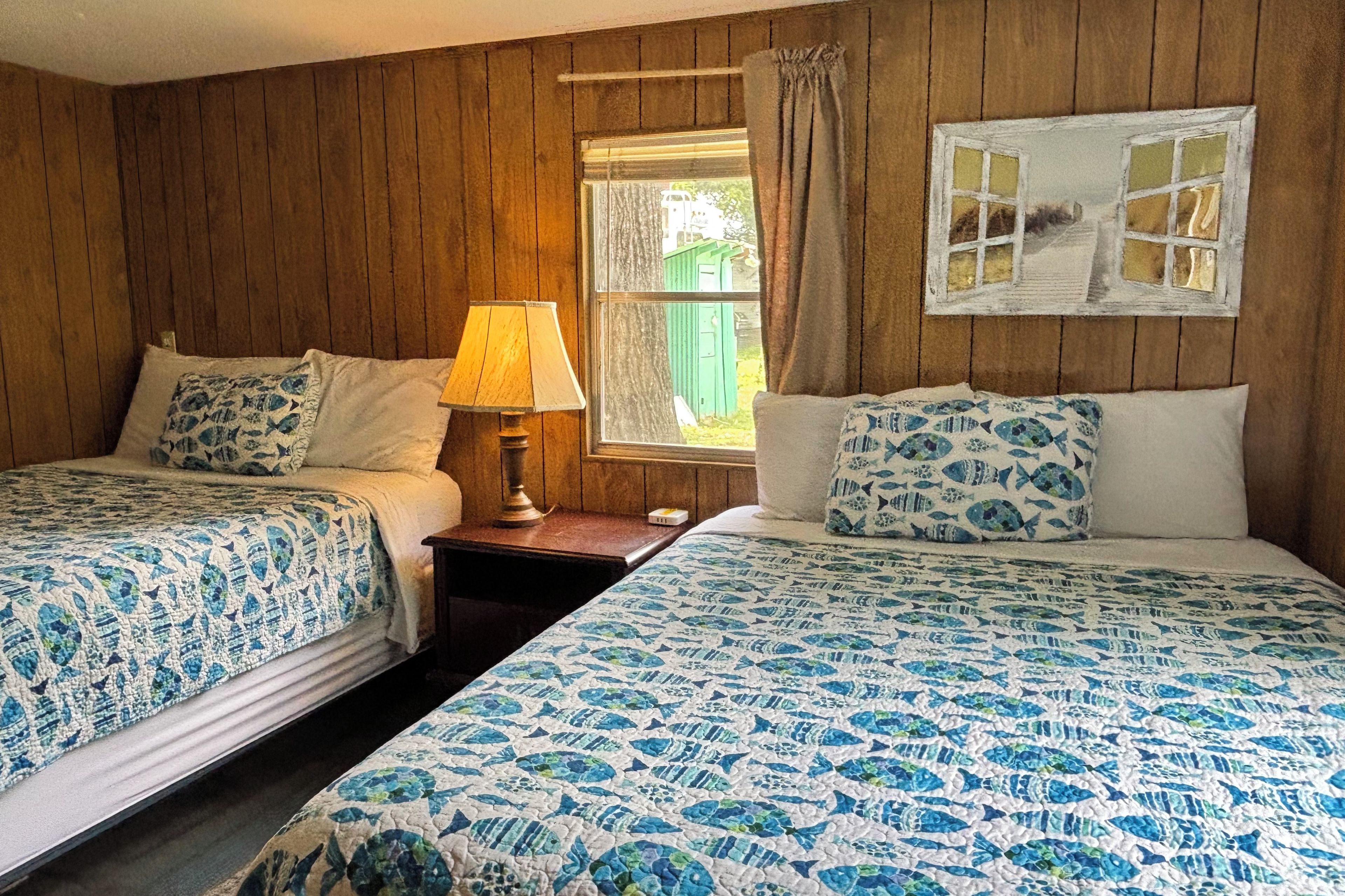 A room with two beds, a nightstand with a lamp, a window with curtains, and a wall-mounted picture.
