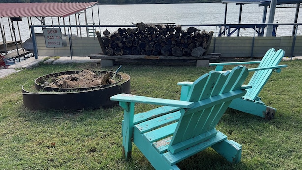 A lakeside deck with a fire pit, stacked firewood, and two turquoise Adirondack chairs.