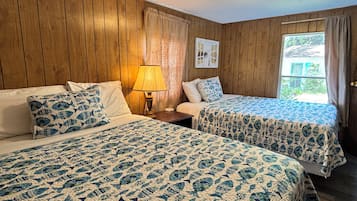 A room with two beds, wooden walls, a window with curtains, a lamp, and a wall art piece.